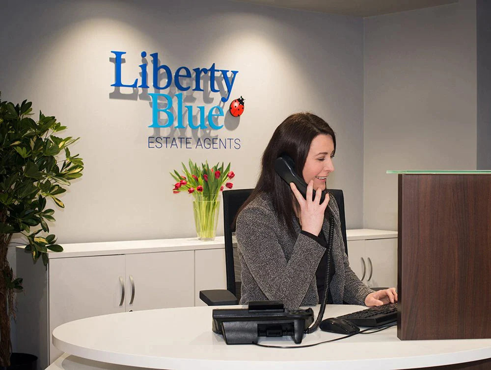 Woman In Grey Talking On Phone In Front Of Liberty Blue Logo